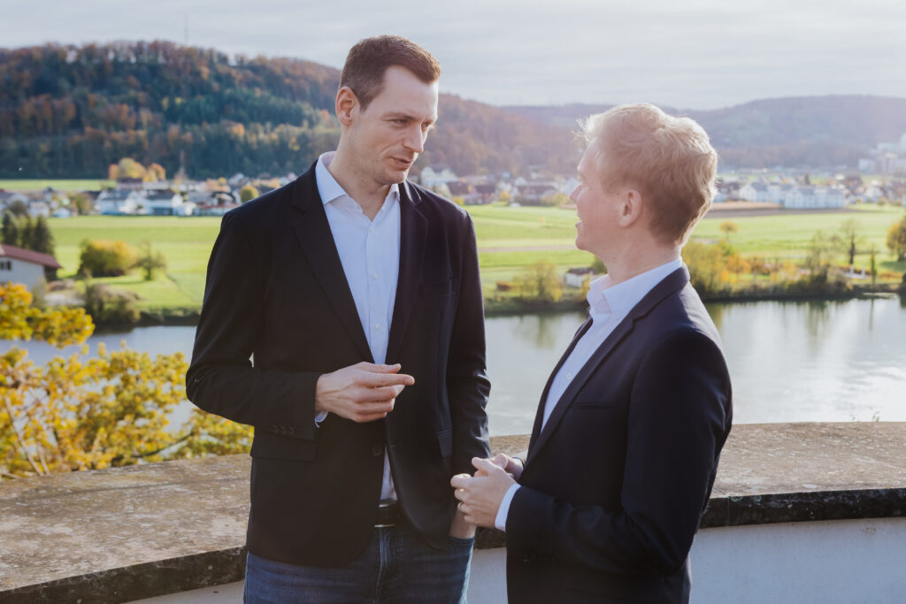 Zwei Männer in Anzügen unterhalten sich mit fließendem Wasser und einer Landschaft im Hintergrund. Herbstfarben sind si...
