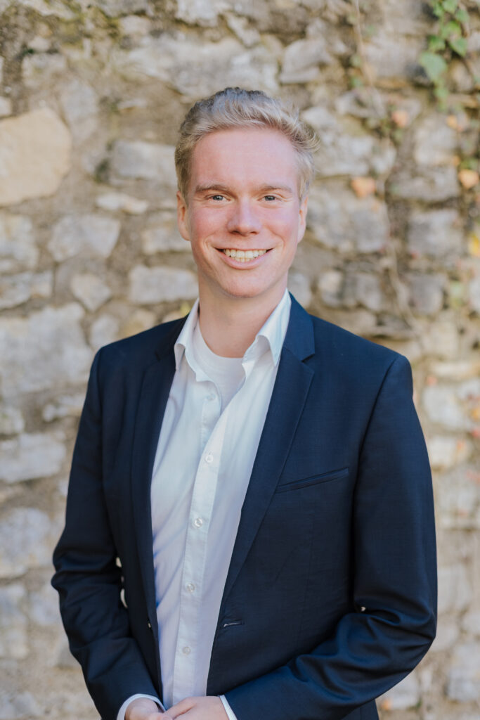Junger Mann mit blonden Haaren in Anzug und weißem Hemd, lächelt vor einer steinernen Mauer.