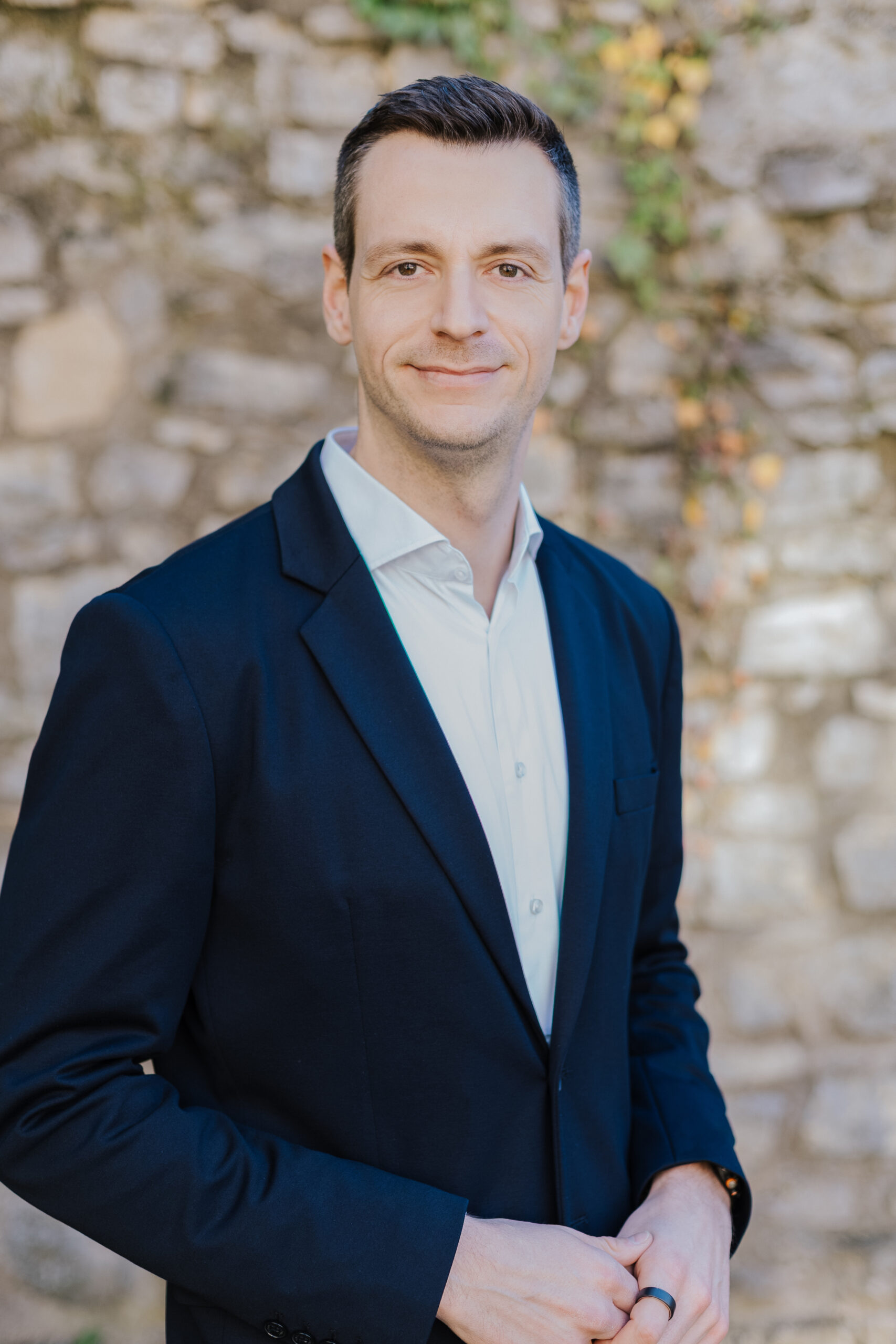 Ein Mann in einem dunklen Anzug lächelt vor einer Steinfassade und hält seine Hände entspannt vor sich.